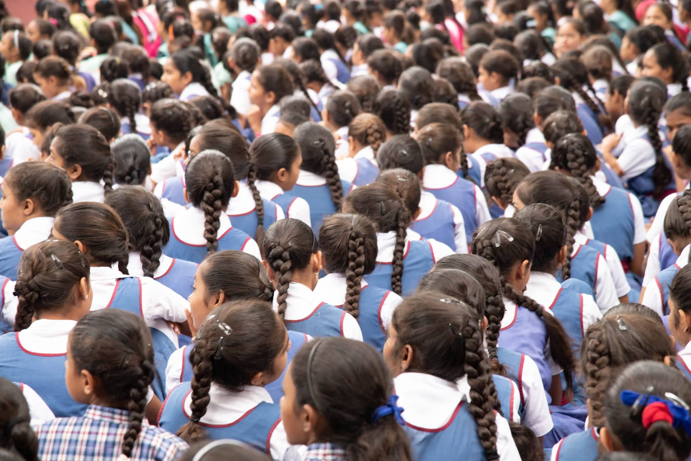 A school assembly of students in uniform seen from behind, seated in rows.